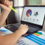 Person analyzing financial charts and graphs on a laptop with colorful documents, showcasing market analysis.